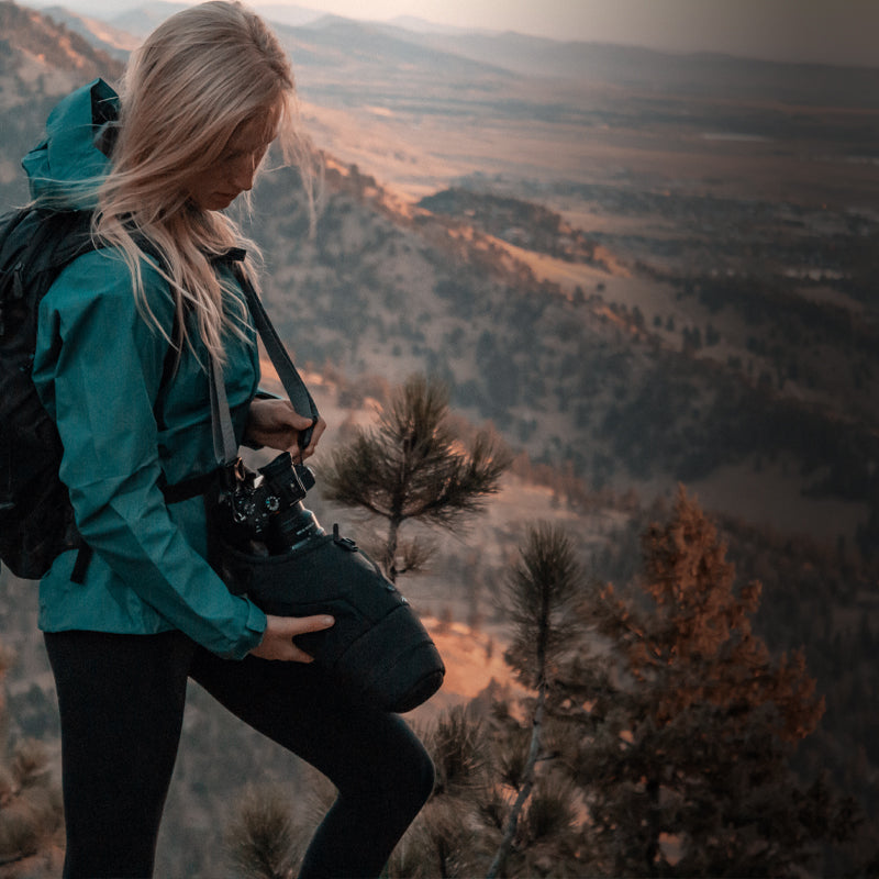 A woman in a teal jacket holds a camera while standing on a mountain, surrounded by trees and a scenic landscape.